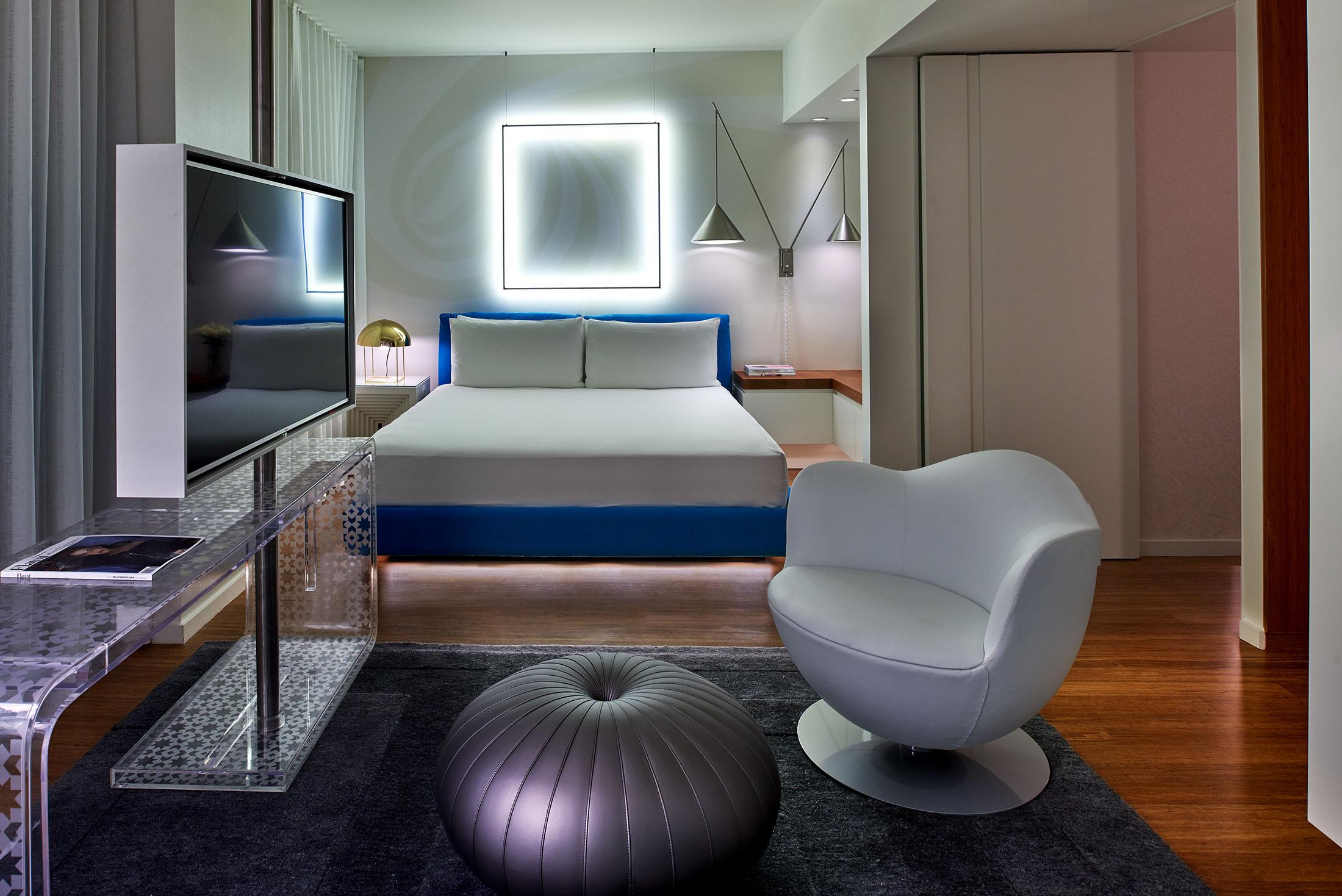 一间旅馆房间，床后有一盏方形灯，前景是一把椅子。